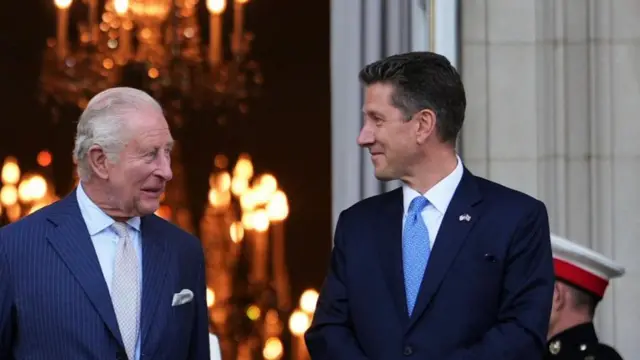 Two men in suits face each other outside an ornately decorated room, seen through a door behind them