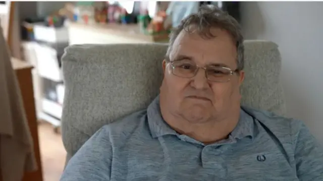 David Chapman wears glasses, a grey polo shirt and has greying short hair. He is sitting back in a chair.