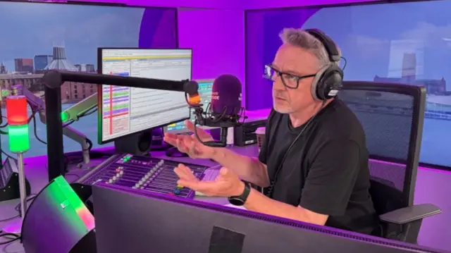 Tony Snell sits behind the microphone in a radio studio, with various pieces of equipment in front of him. He is wearing a black t-shirt, glasses and headphones. He is gesturing with both hands