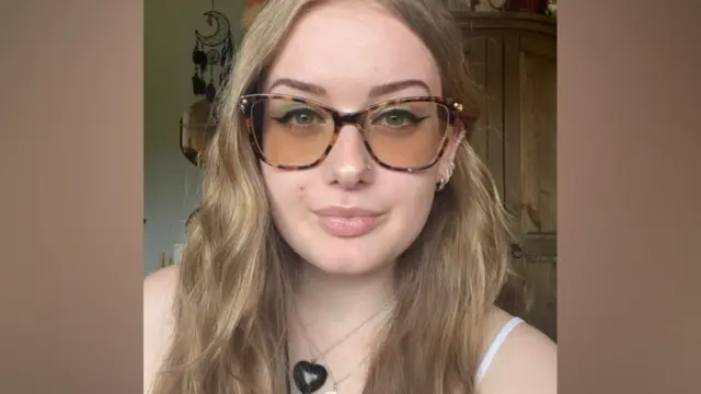 A young woman with long wavy blonde hair and tinted glasses, she has winged eyeliner and a nose piercing.