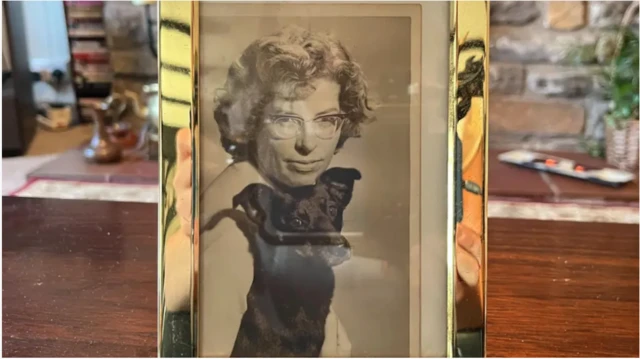 A black and white photo of Iris, holding a small terrier-type dog. It is in a silver frame on top of a wooden piece of furniture and in front of a fireplace.