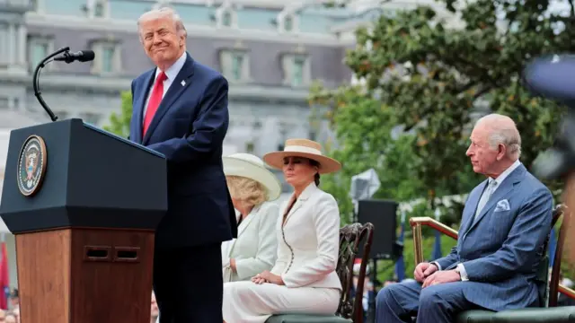 President Trump looks away from the microphone and smiles as he delivers remarks to welcome King Charles III. The King, first lady and queen sit behind him
