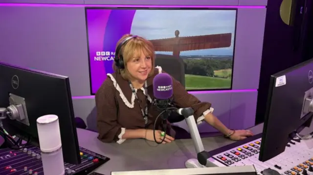 Anna Foster has fair shoulder length hair and is sitting at a radio desk, with a photo of the Angel of the North on a big screen behind her.