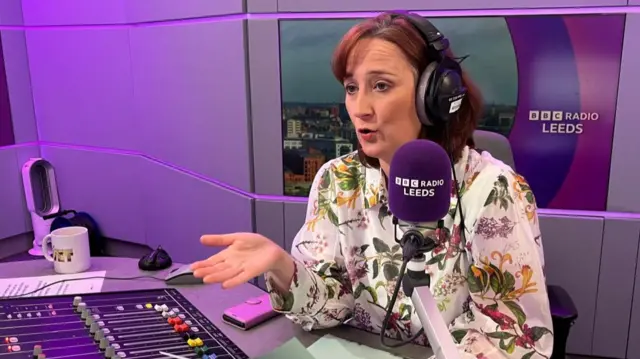 Gayle Lofthouse has reddish shoulder-length hair and is talking into a radio microphone in front of a radio desk.