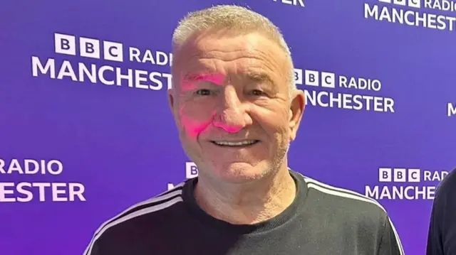 A man with short white hair standing in front of a purple backdrop which says BBC Radio Manchester