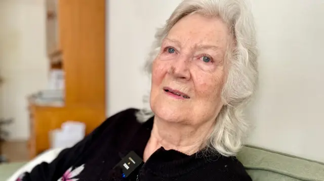 Enid Ford has white shoulder length hair and is relaxing back on a green sofa.