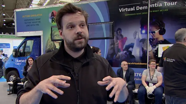 Michael Peachey has short dark brown hair and wears a black top and zip-up jacket. He is holding his hands in front of him as though he is demonstrating.