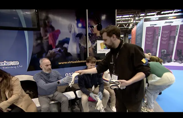 David Gregory-Kumar is wearing a blue top and grey trousers and is passed a pair of gloves while sitting down. He looks on with trepidation.