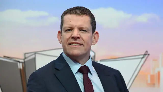Rhun ap Iorwerth has short brown hair. He wears a navy blue suit, with a light blue shirt and maroon tie.