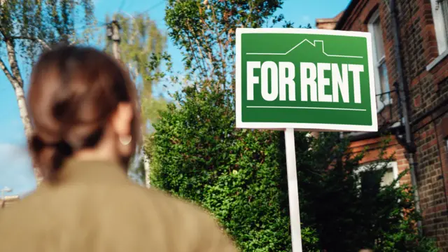 A woman looks at a rent sign outside a house