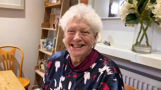 Betty Ruffel, a woman with short, white hair, smiles broadly at the camera, eyes creased up. This is an enormous, animated smile with teeth. She wears a long-sleeved top with splodges of light pink and dark pink on blue and is sitting at what looks like a kitchen table. A shelf to her left has photos on, a pair of glasses is on a shelf behind her and to the right of them, a vase of white flowers.