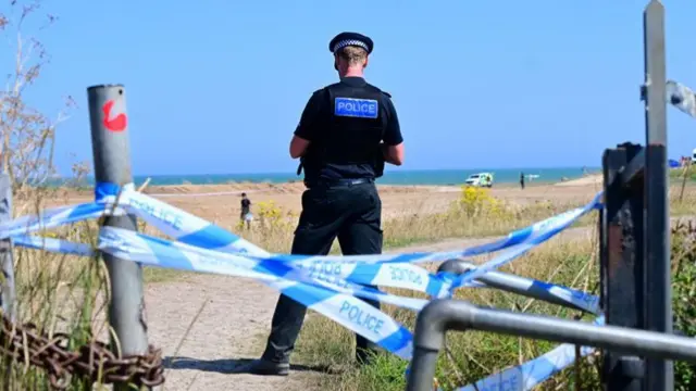 A police officer stands on a beach behind some police tape.