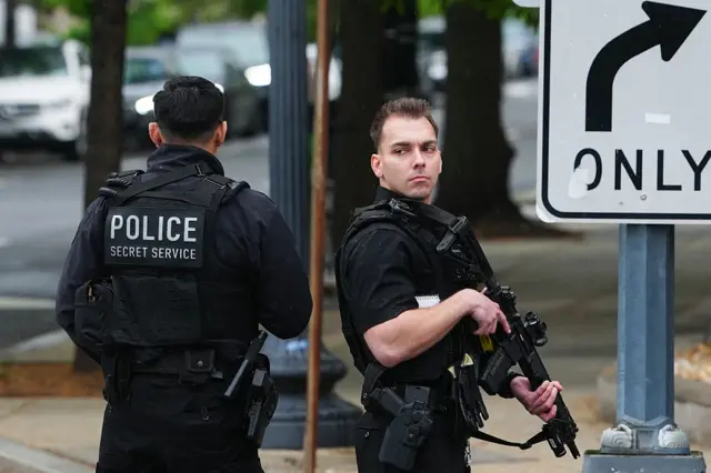 US Secret Service on a road near White House, patrolling with large guns, one facing the camera and the other facing away with 'Police Secret Service' on the back of his uniform