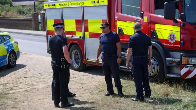 Firefighters and police officers are in a circle. They are talking. Behind them is a fire engine.