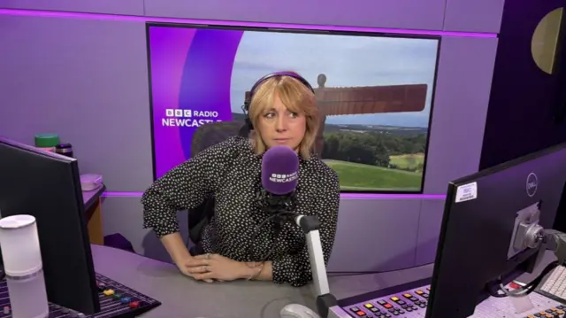 Anna Foster, wearing a polka dot top and headphones, sits behind the microphone in the BBC Radio Newcastle studio, with mixing desks and other equipment in from of her