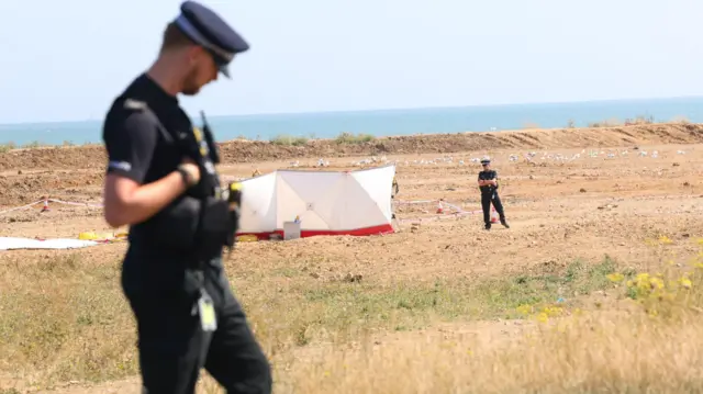 A police officer on a sandy beach. Another police officer is visible in the distance. Between them is a white screen shielding something from public view.