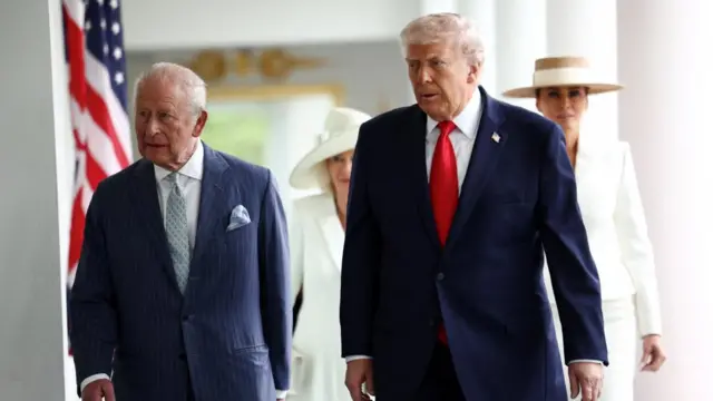 King Charles and Donald Trump walk alongside each other. The King is wearing a navy pinstripe suit and Trump is wearing a navy suit with a bright red tie. Queen Camilla and Melania Trump are blurred in the background.