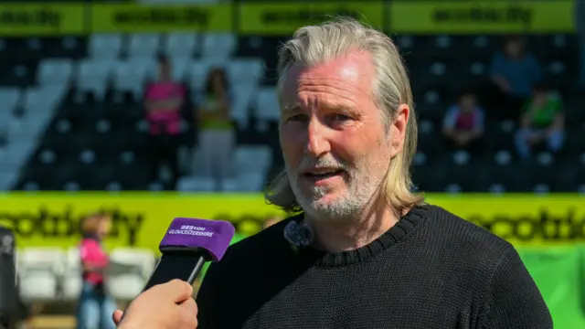Robbie Savage standing inside Forest Green's ground speaking with a BBC Radio Gloucestershire recorder held in front of him