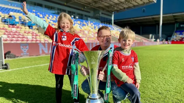 A man in his 30s and two small children in York City football kits hold a large trophy