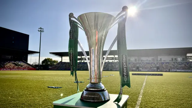 A shiny trophy with ribbons sits on a football pitch