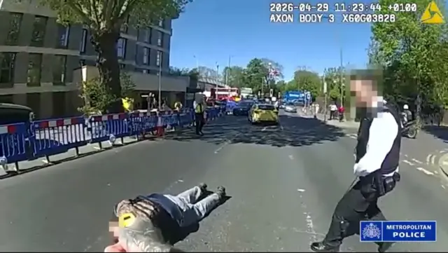 A shot from the body-worn footage of an officer. The suspect is on the ground one officer on the right is approaching him while the hands of the officer whose camera it is can also be seen in the shot