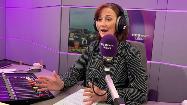 Gayle Lofthouse, wearing headphones and a black and white shirt, gestures with both hands as she talks into the microphone at the BBC Radio Leeds studio, with a mixing desk visible to the left and an image of the city on the wall behind her
