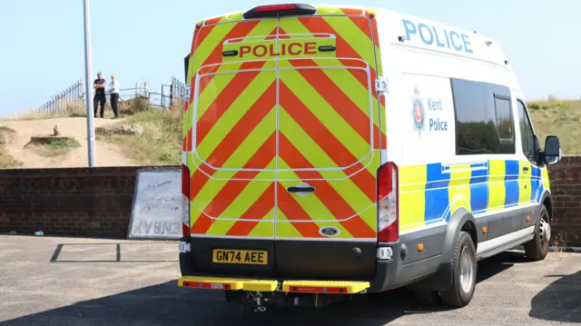 A police van parked in a car park. In the background, police officers stand on a footpath.