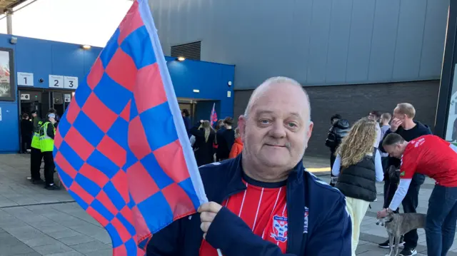 A man wearing a red football shirt and blue jacket hold a red and blue chequered flag