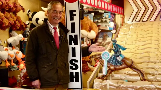 Nigel Farage, wearing a checked shirt, red tie and dark jacket, smiles as he stands by the Finish sign of a fairground attraction featuring racing camels. The winning camel has a Reform UK rosette pinned to it. Prizes including stuff toys can be seen in the background
