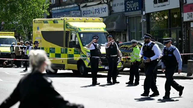 An ambulanec is parked behind a police cordon with multiple police officers standing by it