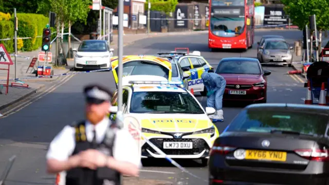 A police officer, out of focus, stands in the foreground of a road block with multiple police vehicles.