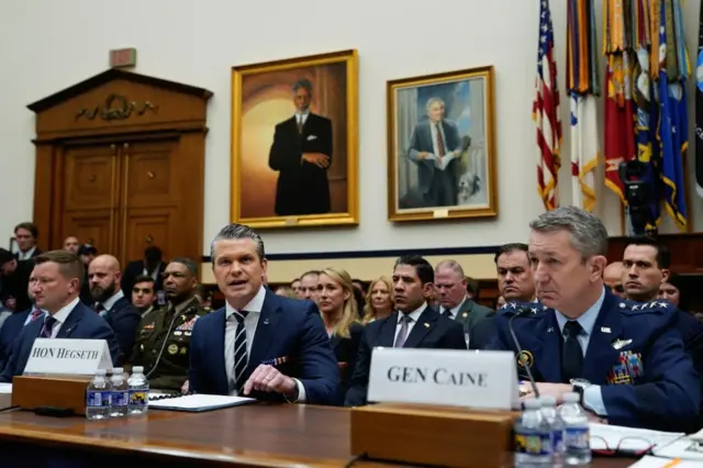 Two men in blue suits and one in a blue military uniform sit at a desk with rows of people sat behind them and portraits and flags adourning the wall