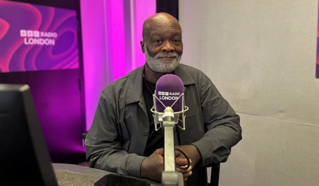 Eddie Nestor sits with his hands clasped behind a purple BBC Radio London microphone in the studio, with a purple screen featuring the station's name in the background