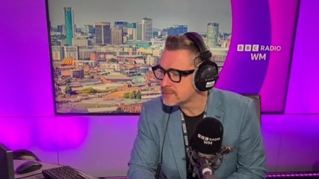 Ed James sits behind the microphone in the BBC Radio WM studio, with an image of Birmingham city centre on the wall behind him. He is wearing thick-rimmed glasses, a black t-shirt and a blue-grey blazer