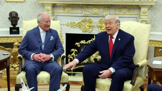 King Charles sits next to Donald Trump in the Oval Office. The King is wearing a navy pinstripe suit and Trump is wearing a dark navy suit with a bright red tie.