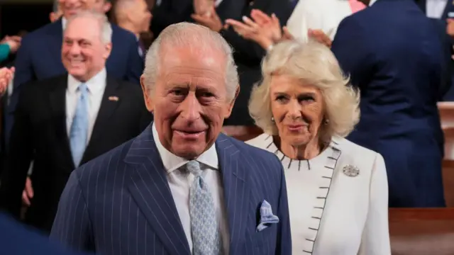 King Charles and Queen Camilla stand in a busy room. The King is wearing a navy pinstripe suit and the Queen is wearing a white jacket.
