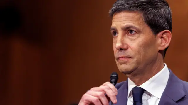 Kevin Warsh holds a slim microphone while sitting in a wood pannelled room