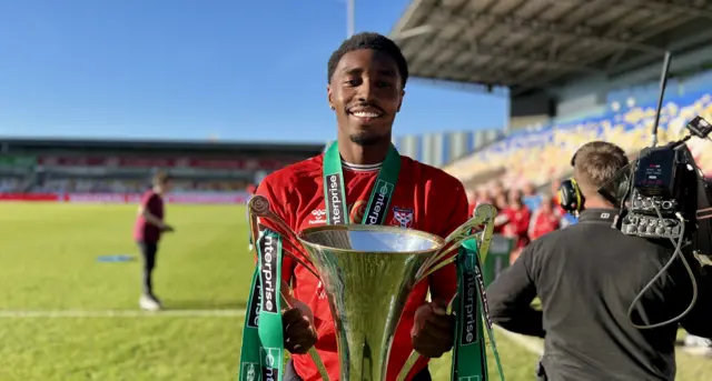 A young man with short black hair wearing a red shirt lifts a trophy