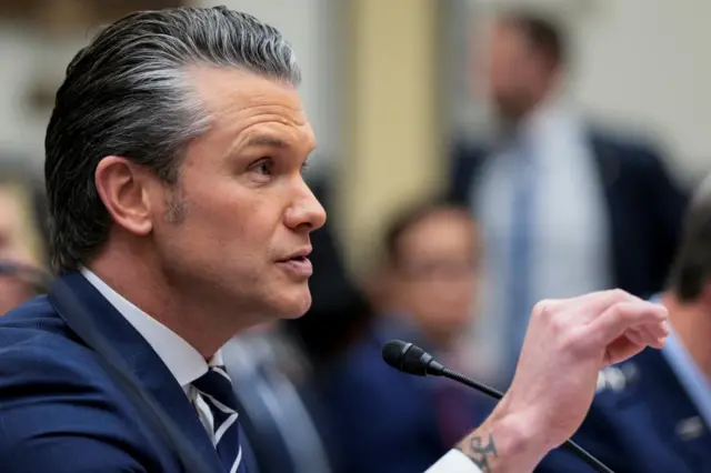 Pete Hegseth, photographed form the side, gestures as he speaks into a microphone