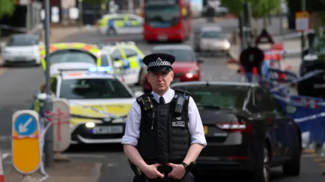 A police officer stands in front of a blocked off street with several vehicles behind him.