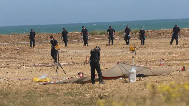 Several police officers comb a beach. Their heads are down and they appear to be looking for things.