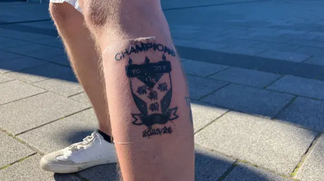 A man's leg with a black tattoo of a football club logo