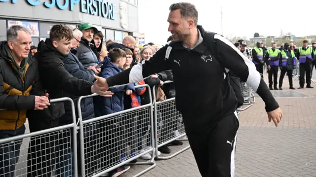Derby County head coach John Eustace, wearing a club tracksuit, smiles and high-fives a fan as he arrives at the CBS Arena to face Coventry