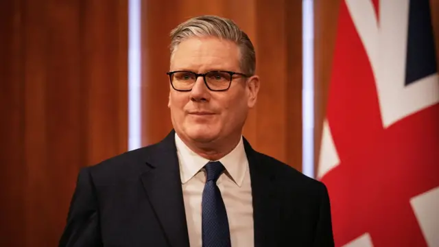Prime Minister Keir Starmer pictured earlier this week wearing a suit and tie with dark-rimmed glasses. A union jack and wood panelling are behind him