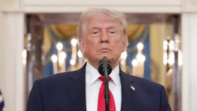 Donald Trump wears a blue suit with a red tie and stands by a microphone