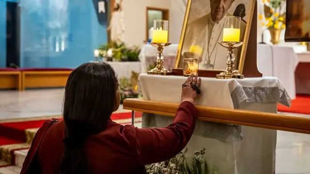 A woman holds a candle up to a photograph of the late Pope Francis