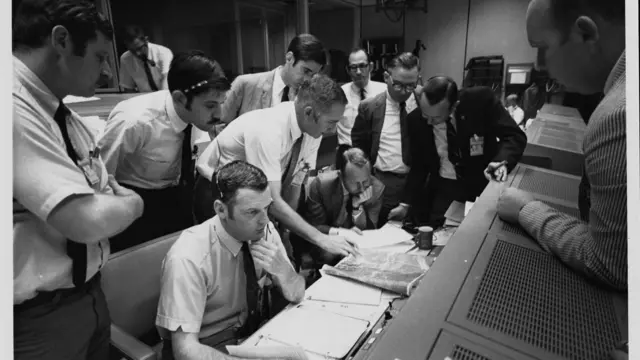 Black and white photo showing a group of flight controllers gather round the console of Shift 4 flight director Glynn Lunney (seated, nearest camera) in the Missions Operations Control Room (MOCR) of the Mission Control Center at the Manned Spacecraft Center in Houston, Texas/