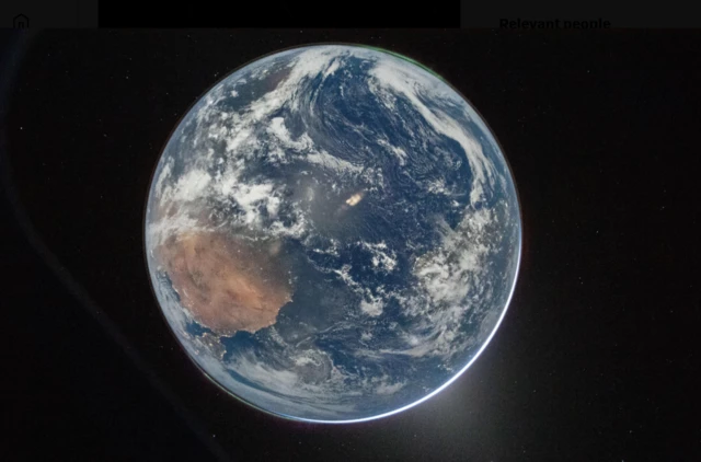 A full disk image of Earth, as seen from the Orion capsule. The planet is a pale blue, swirling with white clouds and glowing slightly lighter blue in place from reflected light. A large brown landmass can be seen on the left hand side