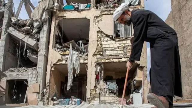 A man in a white turban and black clothes brushes the area in front of a bombed out building