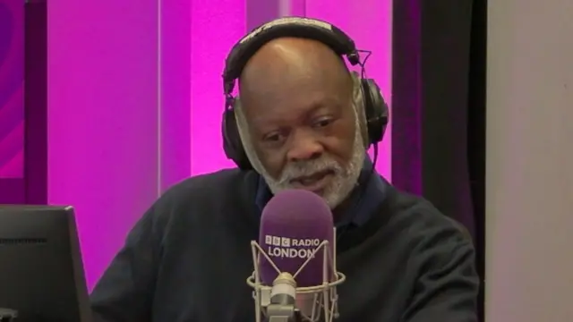 Eddie Nestor looking down to the left of the camera. He is sitting behind a purple BBC Radio London microphone and is wearing black headphones. He is wearing a black jumper. He is sitting in front of a purple background.